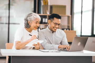 An older mature woman shows something to asian man on a laptop. They smile and work together in an office. They look happy and focused.