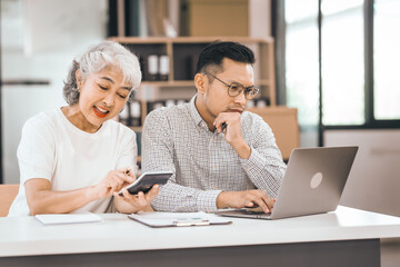 An older mature woman shows something to asian man on a laptop. They smile and work together in an office. They look happy and focused.