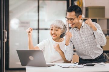 An older mature woman shows something to asian man on a laptop. They smile and work together in an office. They look happy and focused.