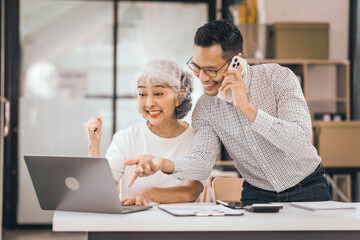 An older mature woman shows something to asian man on a laptop. They smile and work together in an office. They look happy and focused.