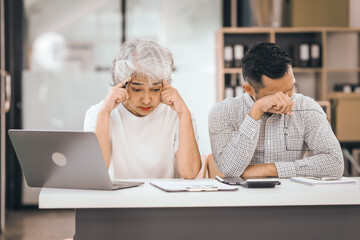 An older mature woman talks to asian man. He covers his face with his hands. They are in an office. They have a laptop and papers. The man looks stressed.