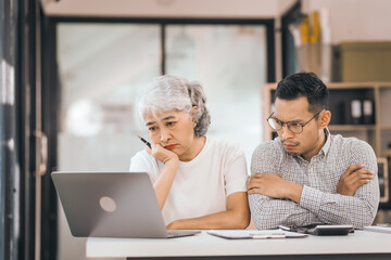 An older mature woman talks to asian man. He covers his face with his hands. They are in an office. They have a laptop and papers. The man looks stressed.