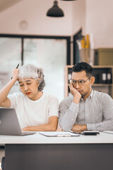 An older mature woman talks to asian man. He covers his face with his hands. They are in an office. They have a laptop and papers. The man looks stressed.
