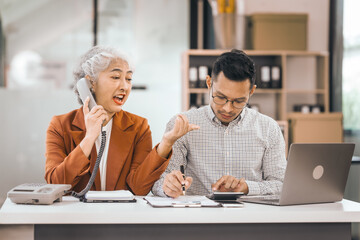 An older mature business woman and asian man work together. She has a tablet. He writes in a book. Both look at their notes. They are focused and happy.
