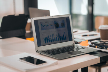 A laptop on a desk displays charts. There's a calculator, coffee, papers, and pens. The workspace seems organized and busy.