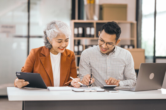 An older mature business woman and asian man work together. She has a tablet. He writes in a book. Both look at their notes. They are focused and happy. - Powered by Adobe