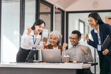 Four Asian business people in office look happy and excited. They raise their fists and smile. sit and stand by table with laptops and coffee. They celebrate together. discuss opinions, presentation