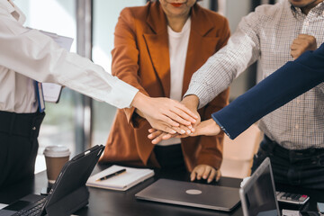 Four people in an office put their hands together as a team. They smile and wear work clothes. laptops, coffee, and papers on the table. They look cooperative. discuss opinions, presentation teamwork