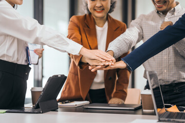 Four people in an office put their hands together as a team. They smile and wear work clothes. laptops, coffee, and papers on the table. They look cooperative. discuss opinions, presentation teamwork