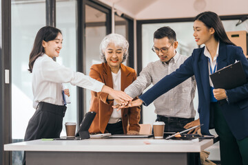 Four people in an office put their hands together as a team. They smile and wear work clothes. laptops, coffee, and papers on the table. They look cooperative. discuss opinions, presentation teamwork