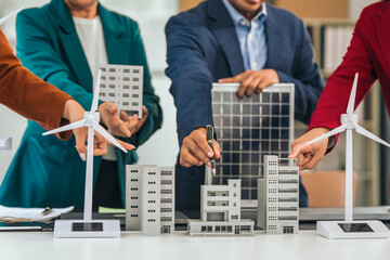 Four asian businesspeople professionals discuss renewable energy in office, showing solar panel and wind turbines with tower building model, apartment. modern business with environmental, clean energy © Phushutter
