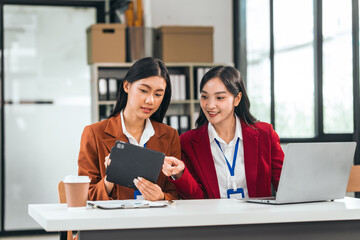 Two young asian business women in the office talk and smile. One holds a tablet, and the other has a pen. They seem excited about their business idea.