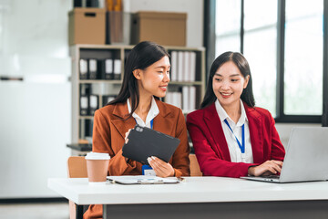 Two young asian business women in the office talk and smile. One holds a tablet, and the other has a pen. They seem excited about their business idea.