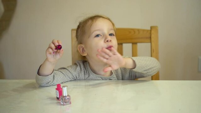 A little girl paints her nails with pink polish. The girl, having put on her makeup, blows on her nails to let them dry.