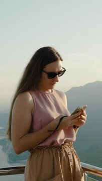 Vertical video. A young woman shares photos of her journey on social media while standing high in the mountains with the sea and sunset in the background.