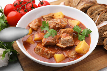 Delicious goulash in bowl, bread and ingredients on white table, closeup