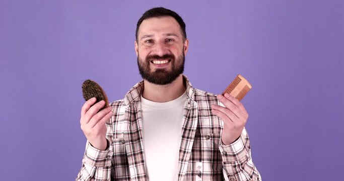 Young bearded man holding comb and brush for beard standing over isolated purple background