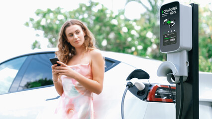 Focused charging station recharging electric vehicle on blurred background of modern woman using...