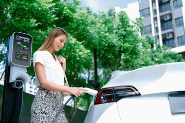 Young woman travel with EV electric car charging in green sustainable city outdoor garden in...
