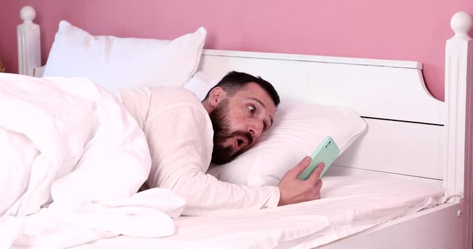 Young bearded man wake up in bed at morning. Young adult taking phone in bedroom. Shocked person looking at phone in bed at morning.