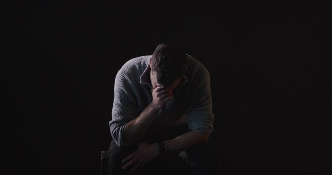 Sitting, sad man sobs in dark room, black background. Static