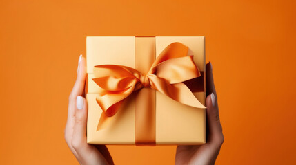 A woman's hands hold an orange gift box with a bow and ribbons on a colorful background - obrazy, fototapety, plakaty