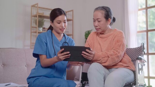 young Asian female nurse, sitting on the sofa, holds a tablet and provides a detailed explanation to an elderly Asian lady in a wheelchair in the living room.