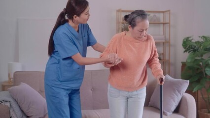A young Asian female caregiver is assisting an elderly Asian woman, supporting her with a walking cane as they sit together on the sofa in the living room.