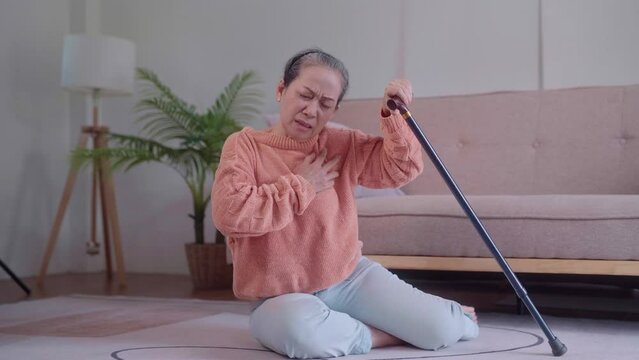 An elderly Asian woman, holding a walking cane, attempts to rise from the sofa but falls back, possibly due to a heart-related issue, in the living room. Her need for assistance is evident.
