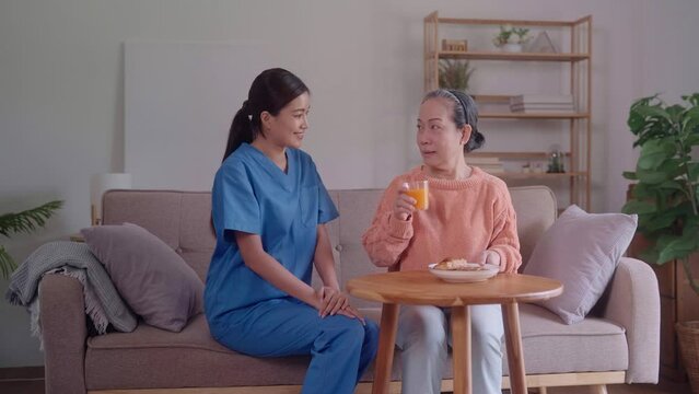 Asian female caregiver serves toasted bread and fruit juice to an elderly Asian woman, sitting on a sofa in the living room. This image portrays the attentive care and kindness provided to seniors.