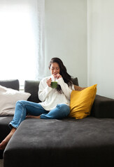 Healthy lifestyle. Young Chinese woman drinking green vegetable juice at home sitting on the sofa. Vertical image.