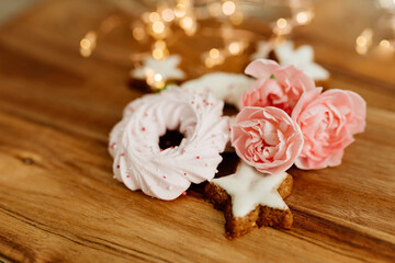 Gingerbread cookies in the shape of stars, merengue cookies and pink roses on a wooden table against a background of bokeh of New Year’s lights. Christmas background. Winter card. Delicious pastries