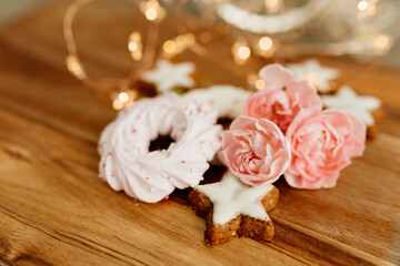 Gingerbread cookies in the shape of stars, merengue cookies and pink roses on a wooden table against a background of bokeh of New Year’s lights. Christmas background. Winter card. Delicious pastries