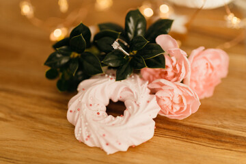 Pink merengue cookies and pink roses on a wooden brown natural table against a background of bokeh of New Year’s golden lights. Engagement ring with diamonds. Christmas background. Valentine’s Day