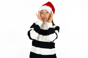 Beautiful girl wearing striped sweater and Santa hat with scared expression, keeps hands on head, jaw dropped, has terrific expression. Omg concept
