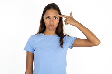 Unhappy Beautiful girl wearing blue T-shirt makes suicide gesture and imitates gun with hand, curves lips keeps two fingers on temple, shoots, being tired of everything,