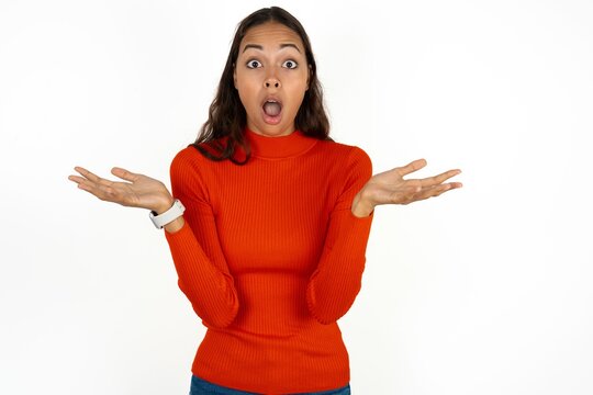 Frustrated Beautiful girl wearing red sweater feels puzzled and hesitant, shrugs shoulders in bewilderment, keeps mouth widely opened, doesn't know what to do.