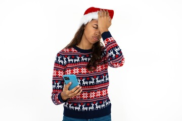 Upset depressed Beautiful girl wearing christmas sweater and Santa hat makes face palm as forgot about something important holds mobile phone expresses sorrow and regret blames