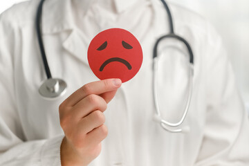 Close up of young female doctor in white uniform with stethoscope holding red angry face emoticon....
