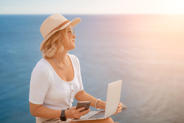 Freelance women sea working on the computer. Good looking middle aged woman typing on a laptop keyboard outdoors with a beautiful sea view. The concept of remote work.