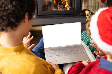 Diverse gay male couple wearing christmas hats and having laptop video call at home, copy space
