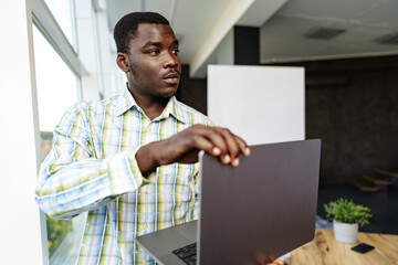 Young african man standing near the window indoors and holding laptop