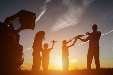 Black silhouettes of family members fly kite near car with open trunk in evening. Concept of...