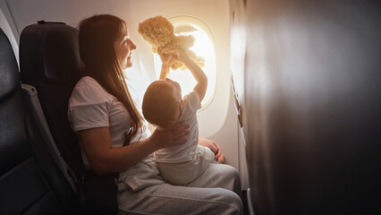 Mom takes flight and cradles toddler girl in arms. Toddler daughter joyfully plays and raises up toy bear during journey, sunlight - obrazy, fototapety, plakaty