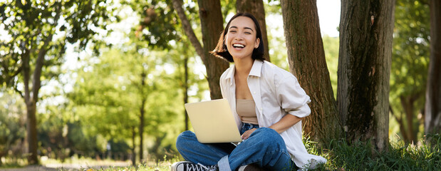 Portrait of asian girl with laptop, sitting near tree in park on sunny summer day, working on...