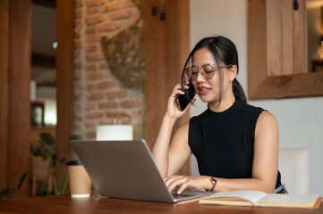 A professional Asian businesswoman is talking on the phone while working remotely from a coffee shop