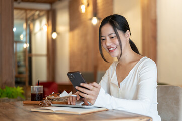 A happy Asian woman is using her smartphone, texting her friends while sitting in a coffee shop.