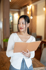 A beautiful Asian woman in casual clothes holding a book while standing in a beautiful coffee shop.