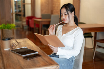 A thoughtful and focused Asian woman is reading a book and working remotely at a cafe.