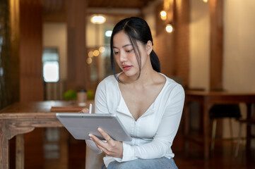 A beautiful and focused Asian woman is using her digital tablet while sitting in a coffee shop.
